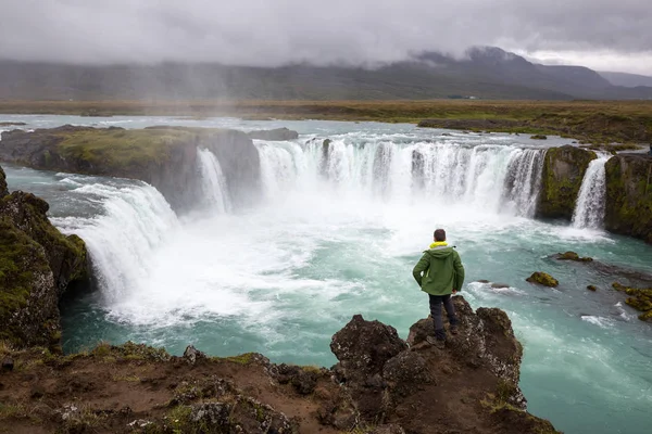 Um turista masculino fica em um penhasco acima de uma cachoeira e admira sua beleza. Fotografia tirada na Islândia . — Fotografia de Stock