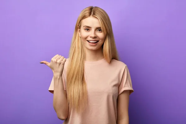 Optimistic young girl with toothy smile points aside with thumb — Stock Photo, Image