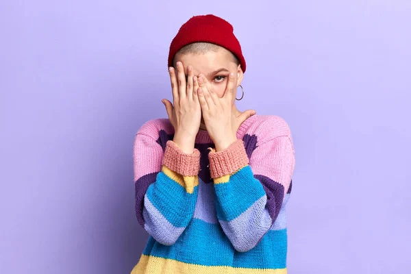 Застенчивая крутая гламурная девушка в полосатом разноцветном свитере закрывает лицо руками — стоковое фото
