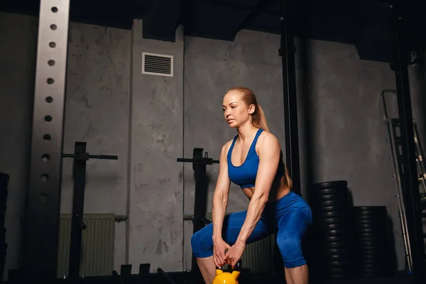 fair-haired woman in stylish blue sportwear doing squat with yellow weights