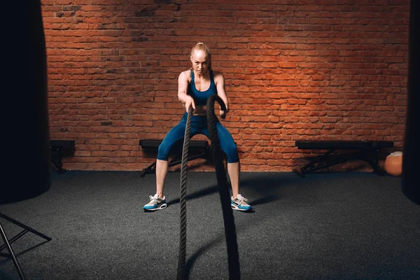 fair-haired beautiful serious athletic woman doing battle rope exercises at gym