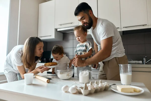 girl smelling yummy hot pancakes while elder sister and her daddy mixing dough