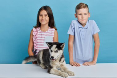 Utangaç küçük çocuklar bir köpekle masanın arkasında duruyorlar.
