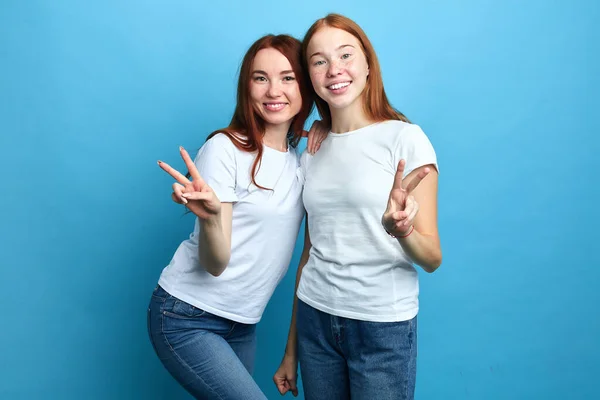 Usmívající se dobře vypadající sestry v bílých tričkách a džínách ukazuje vítězství gesto — Stock fotografie