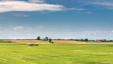 Mavi gökyüzü, sarı alan ve traktörlerin çalıştığı yeşil çayırlı peyzaj.
