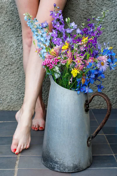 Bouquet of multicolored wildflowers in an old metal jug against the background of female legs. — Stock Photo, Image