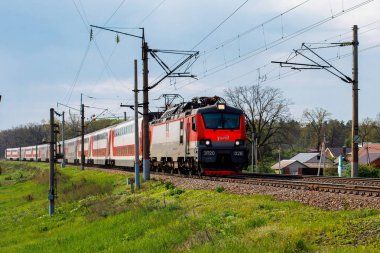 Voronezh, Rusya: 14 Mayıs 2019. Bir elektrikli lokomotif sero-kırmızı bir yolcu treni çeker. Ön planda, yeşil çim, arka planda Kuru ağaçlar.