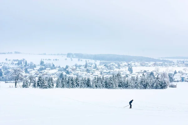 Paesaggio invernale con alberi ghiacciati e un piccolo villaggio europeo situato tra campi innevati. Sciatore nel mezzo di un campo innevato — Foto Stock