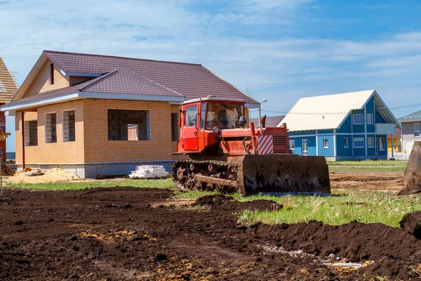 An old red crawler tractor spreads the ground near unfinished small houses. — Stock Photo, Image