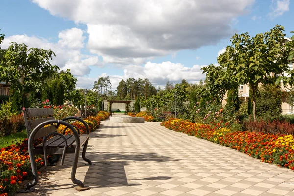 Mejora de la aldea rural. En primer plano hay un banco de madera. La pasarela pavimentada conduce a lo largo de macizos de flores, arbustos ornamentales y árboles en un moderno pueblo rural en un día soleado de verano. . — Foto de Stock