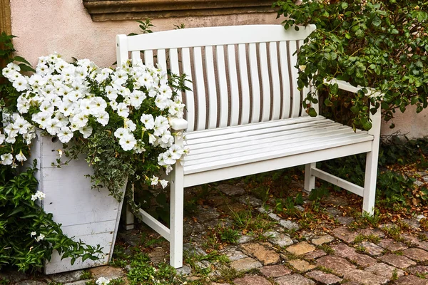 Sui ciottoli vicino ai fiori di petunia bianca che crescono in una grande scatola di legno si trova una comoda panca di legno bianca. Il comfort di una piccola città . — Foto Stock