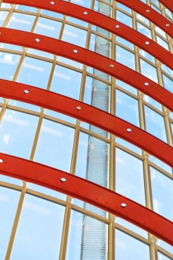 Kavisli kırmızı ve beyaz metal yapılar pencere sistemlerini tutar. Camın içinden parlak güneş ışığı geçiyor. Demir yapının gücü ve havasının yansıması..