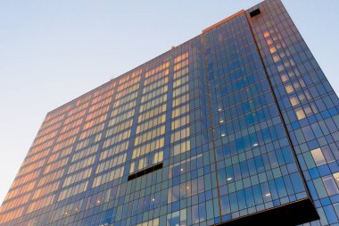 Günbatımında çok katlı bir binanın cam cephesi, pencereler güneşin turuncu ışığını yansıtır. Kentsellik temasının tasarımı için.