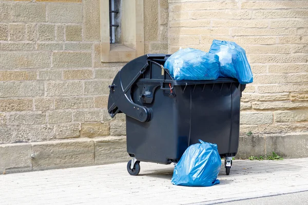Black plastic dumpster with full blue trash bags on the street — Stock Photo, Image