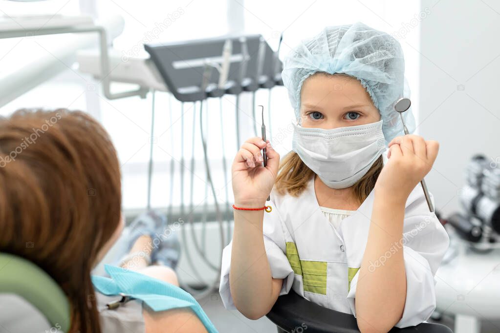 Caucasian seven year old girl playing at the dentist. Child holding dental instruments in hands, woman dentist depicts a patient