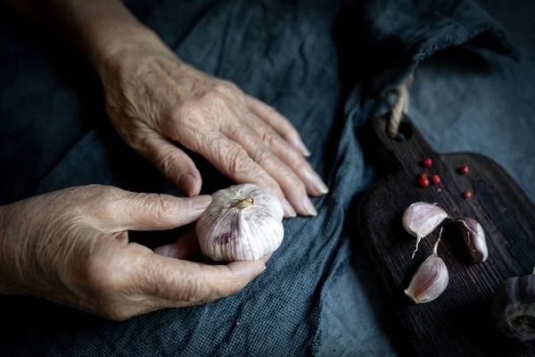 Mãos de mulher sênior com uma cabeça de alho em um fundo têxtil cinza. Close-up, foco seletivo — Fotografia de Stock
