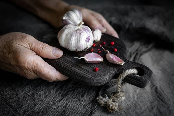 Elderly womans hands hold a chopping board with garlic over a grey textile background. Close-Up, selective focus Stock Photo