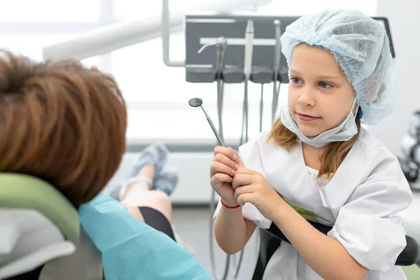 Kaukasische Siebenjährige tauschte mit ihrem Zahnarzt in einer Klinik oder Praxis die Rollen. Kind trägt Arztuniform, Schutzmaske und Zahnspiegel — Stockfoto