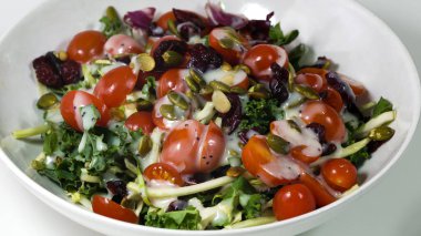 Yeşil yapraklı sebze salatası karışımı, domates, kabak çekirdeği, kurutulmuş kızılcık ve yakın bir tabakta sos..