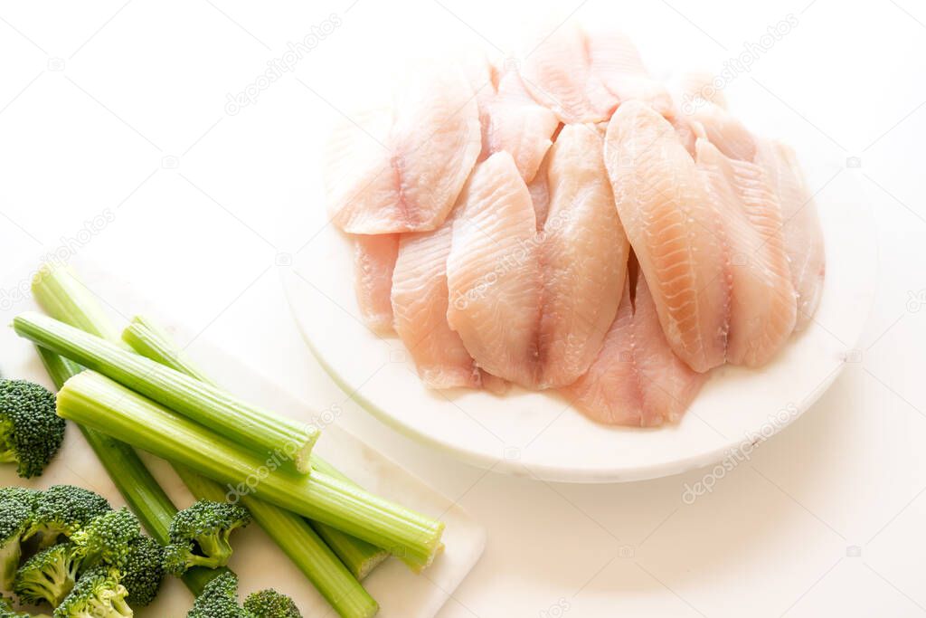 Raw Tilapia fish fillet and fresh vegatables, broccoli and celery closeup on white background