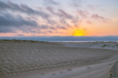 Gün batımında sahilde kum tepeleri. Arkaplanda güneş ufuk çizgisinde ve bulutlu gökyüzünde batıyor