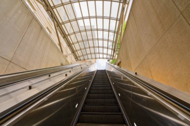 Modern cam çatılı ve çevreleyen beton duvarlı bir metro istasyonundan yükselen bir yürüyen merdiven manzarası..