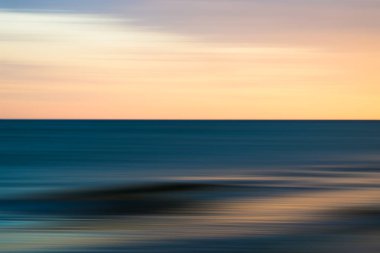 Canlı bir günbatımı gökyüzünün altında, hareket bulanıklığı içinde bir dalga kıyıya doğru yuvarlanıyor..