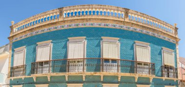 Kapalı panjurları, çiçekli süslemeleri ve işlenmiş demir balkonları olan mavi kiremitlerle kaplı klasik bir binanın kıvrımlı ön cephesi..