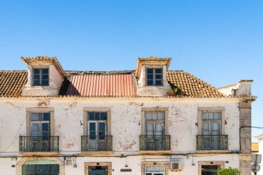 Açık mavi gökyüzünün altında, kabuğu soyulmuş, kiremitli çatısı ve demir balkonları olan eski, beyaz boyanmış bir bina..
