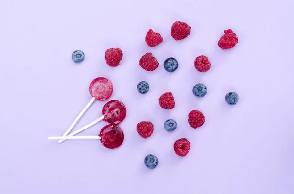 Piruletas rojas, arándanos y frambuesas sobre un fondo de color violeta. Vista superior plano Lay fondo . —  Fotos de Stock