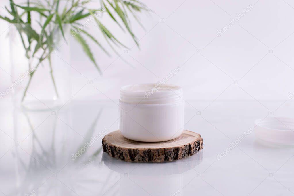 Natural cosmetics with cannabis oil. Mockup with a White clean jar with cosmetics cream and fresh green leaves of hemp.