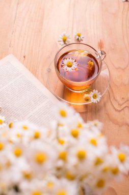 Papatya çiçekli bir fincan sıcak bitki çayı ahşap bir masada kitabı açtı. Sabah kahvaltısı kavramı.