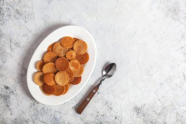 Modaya uygun ev yapımı kahvaltı, krep gevreği, beyaz tabakta mini krepler, kaşıkla beyaz beton arka plan.