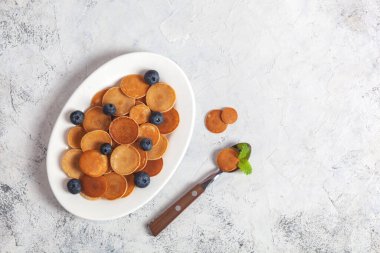 Modaya uygun ev yapımı kahvaltı, krep gevreği, beyaz tabakta mini krep, beyaz beton arka planda kaşık ve yabanmersini, limon merhemi ve nane yaprakları.