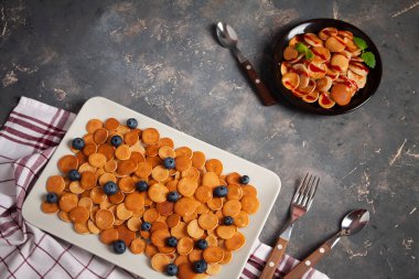 Modaya uygun ev yapımı kahvaltı, krep gevreği, tabaklar üzerinde mini krepler koyu beton arka planda kaşık ve yabanmersinli, nane ve tekstil.