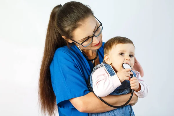 Lékařka mladá žena drží dítě v náručí a vypadá na to, a dítě si hraje s stetoskop. — Stock fotografie