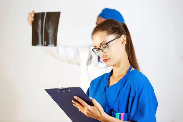 En el fondo, el médico observa una radiografía, en primer plano una doctora escribe información en un diario. Fondo blanco — Foto de Stock