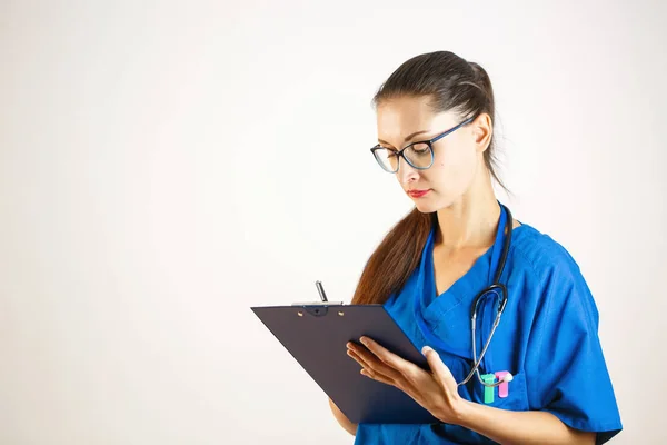 Una doctora con una bata azul está registrando datos en un diario, un estetoscopio está en su cuello. Fondo blanco — Foto de Stock