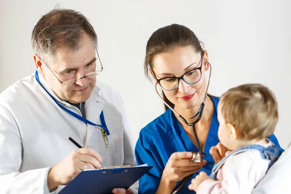 Dos médicos examinan a un niño pequeño, uno usando un estetoscopio y el otro tomando notas en una tarjeta — Foto de Stock