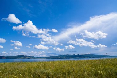 Çiçekli bozkır ve Sibirya nehri üzerinde bulutlarla kaplı mavi bir gökyüzü. Arka planda dağlar. Krasnoyarsk bölgesi