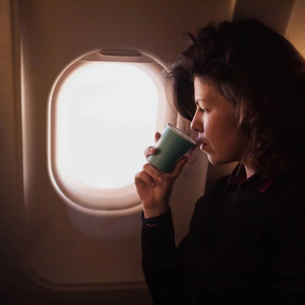 Young woman drinking coffee inside airplane while sitting on passenger seat near window