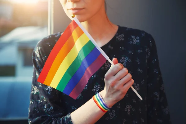 young woman with rainbow ribbon wristband  on her hand holding lgbt colorful rainbow flag next to her face