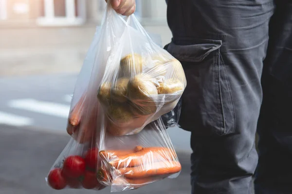 man's hand holding plastic bags with fresh vegetables from farmer's market, walking on city street