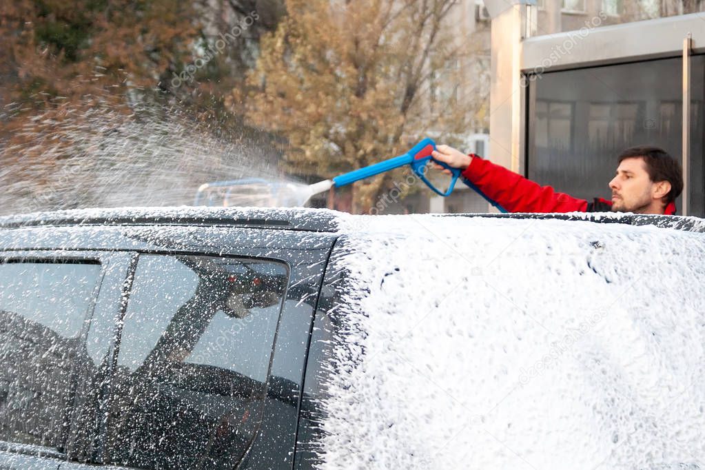 A man in a red jacket covers a black car with foam at a self-service car wash. Back view