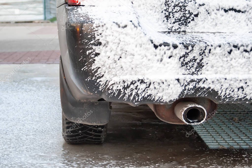 Black car being covered in foam on the self-service car wash. Close-up on the rear bumper, headlight, exhaust pipe and wheel