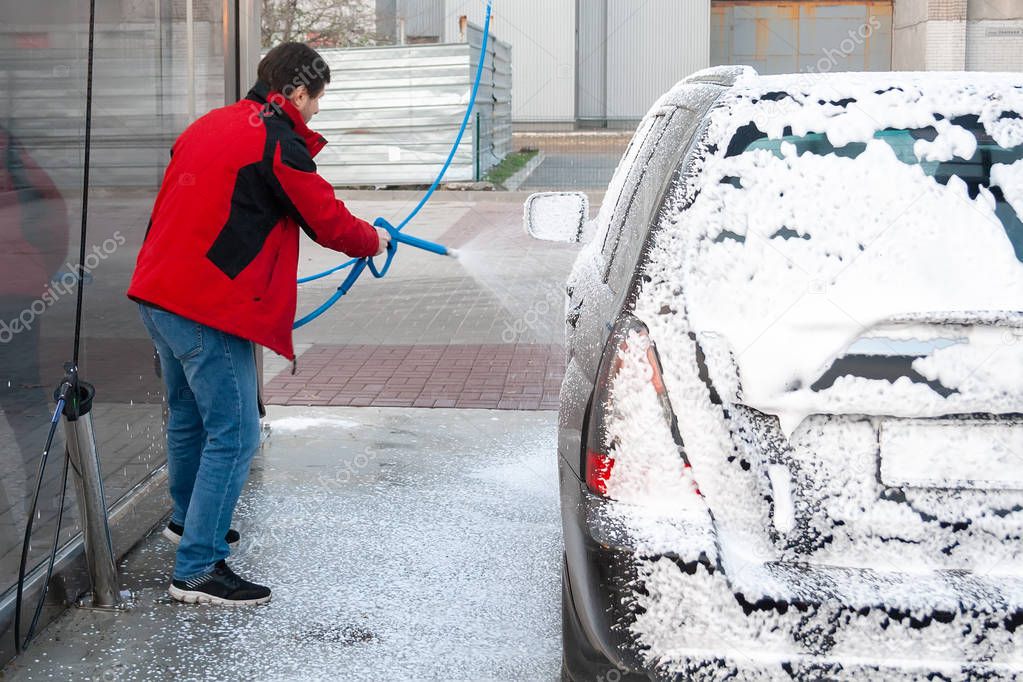 A man in a red jacket covers a black car with foam at a self-service car wash. Back view