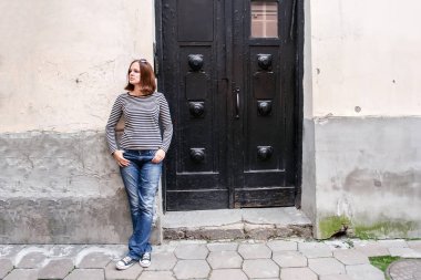Bir kız kot pantolon, çizgili bir kazak ve spor ayakkabı içinde eski bir ev, siyah ahşap kapının yanında duran ve yan görünüyor