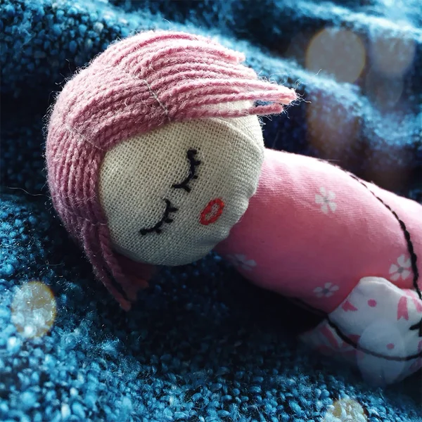 Little sleeping handmade doll made of linen and cotton lying on a blue plaid. She has pink hair, closed eyes, a flowered dress. She holds a pillow in her hands from the threads — Stock Photo, Image