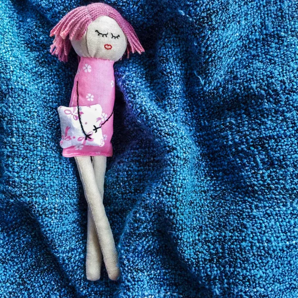 Little sleeping handmade doll made of linen and cotton lying with crossed legs on a blue plaid. She has pink hair, closed eyes, a flowered dress. She holds a pillow in her hands from the threads — Stock Photo, Image