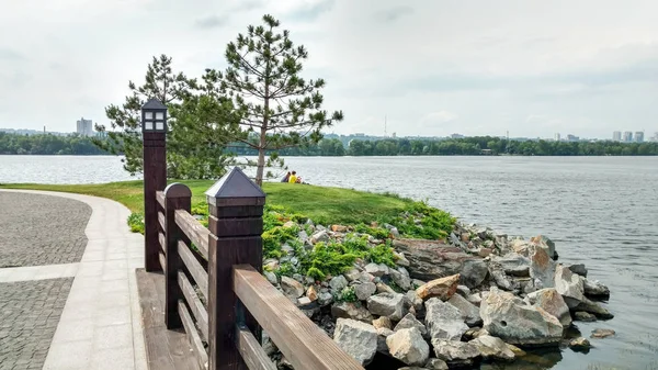 Vista panorámica desde un pequeño puente de madera a un río ancho y una pequeña isla con una orilla de piedra, césped cuidadosamente recortado, pinos y una pareja descansando a distancia —  Fotos de Stock